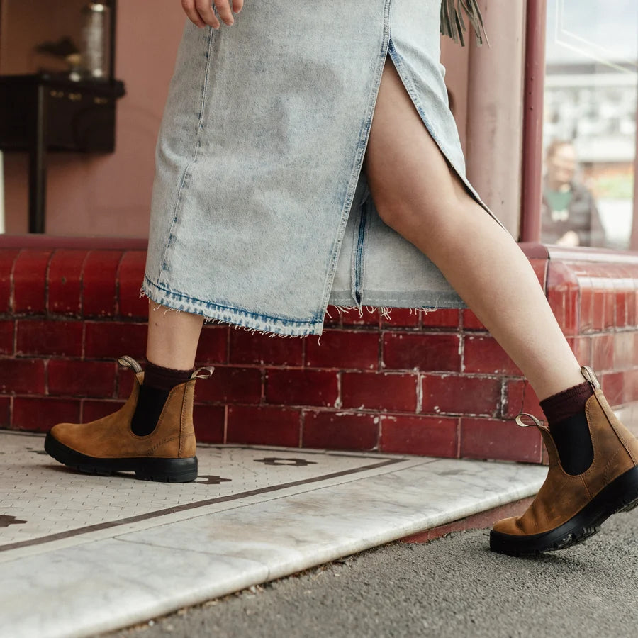 A person wearing a light blue denim skirt and brown Chelsea-style ankle boots with black elastic side panels, standing on a sidewalk beside a red brick wall.